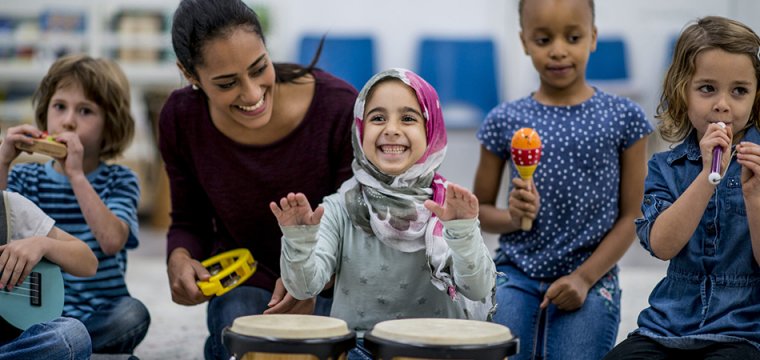 Coram charity image showing a multi-ethnic group of young children in a classroom. Their teacher is watching them playing instruments together. The instruments include drums, maracas, and a guitar.